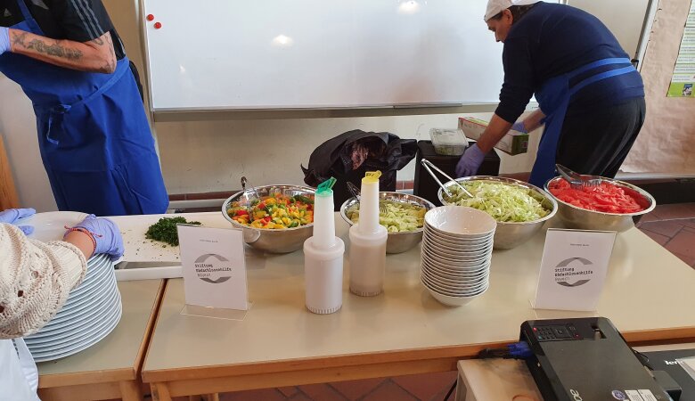 Essensausgabe auf dem Tisch stehen Teller Schüssel mit Salat dahinter stehen zwei Helfer die alles vorbereiten