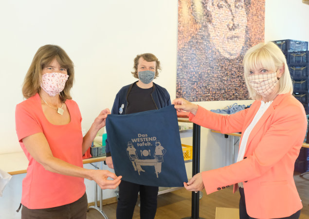 zwei Mitarbeiter mit der Sozialministerin Carolina Trautner präsentieren Einkaufsbeutel mit dem Logo des Projekts